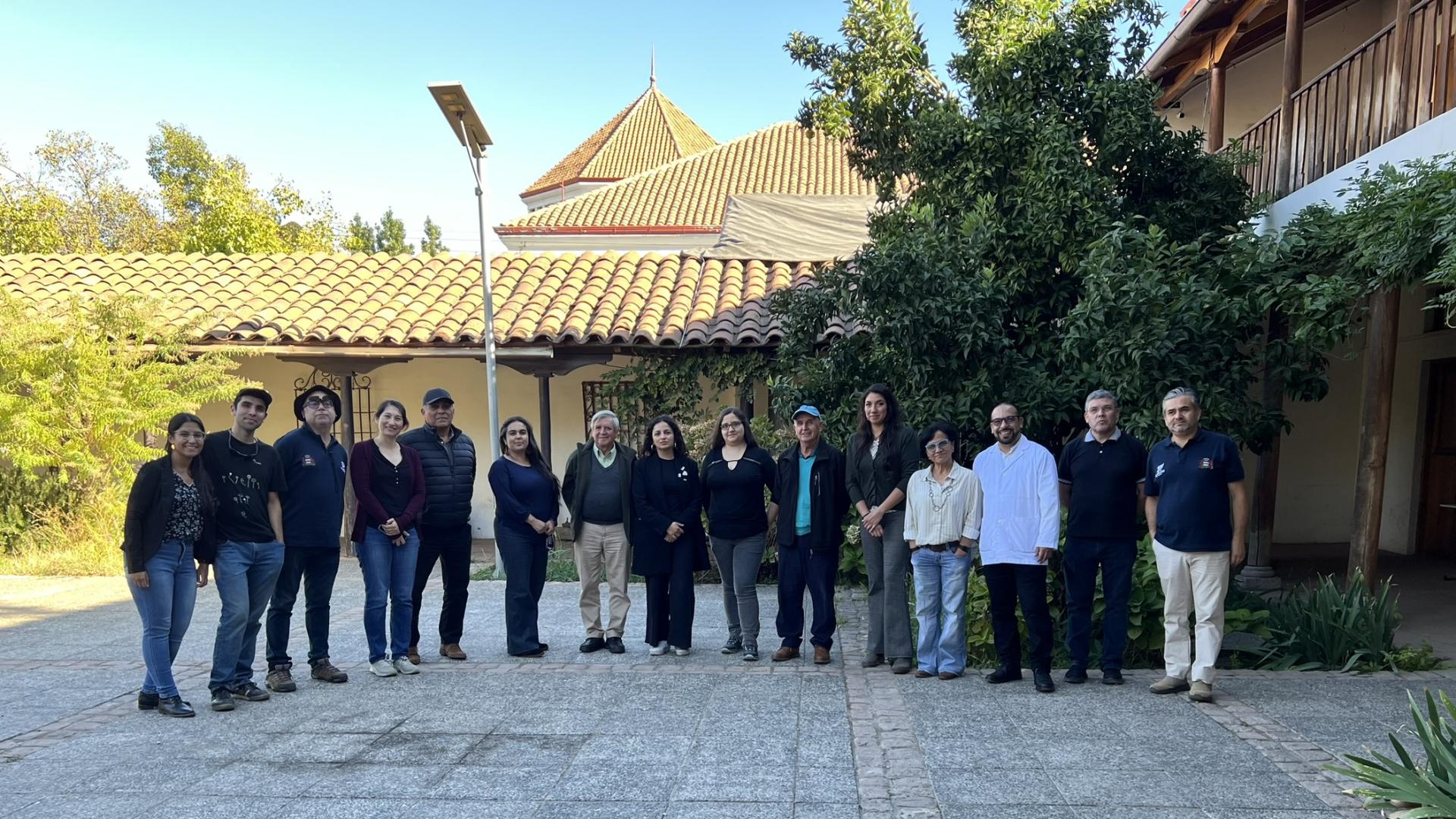 Fotografía en que se aprecian 15 personas adultas en fila, posando hacia la foto. De fondo hay algunos árboles y una planta de una casa colonial, murallas blancas de adobe y techos de teja en tonos marrón. 