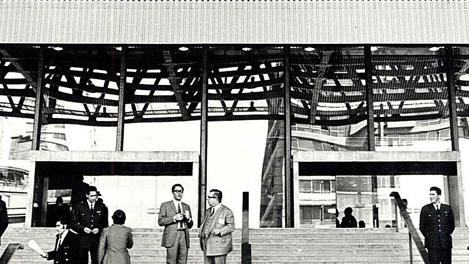 Imagen en blanco y negro de grandes escaleres con gente del frontis de un edificio y con techo de  vitrales.