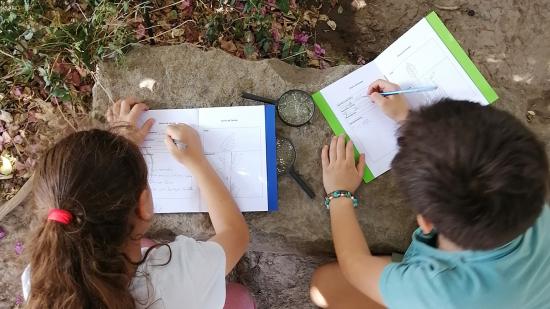 Niña y niño escriben en un cuadernillo sobre una roca. 
